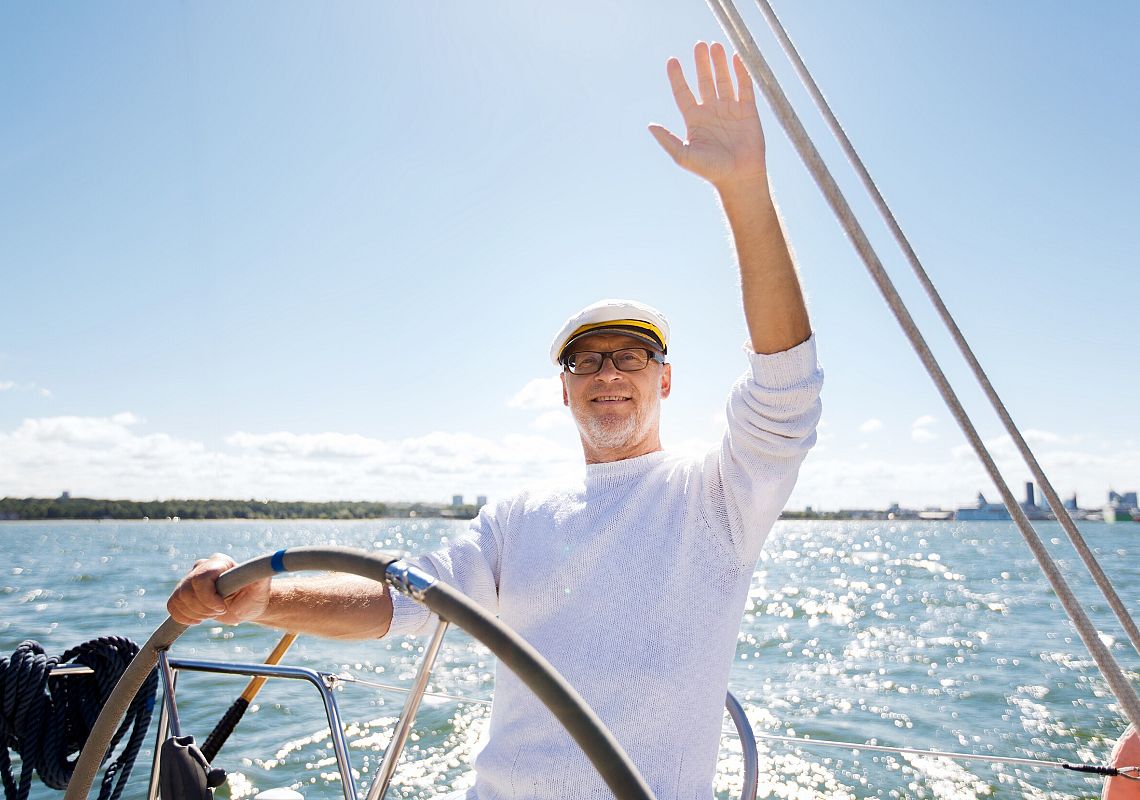 Imprezy Zorganizowane – Mężczyzna w średnim wieku, w stroju żeglarskim, stojący za steram jachtu, macha w stronę kamery.